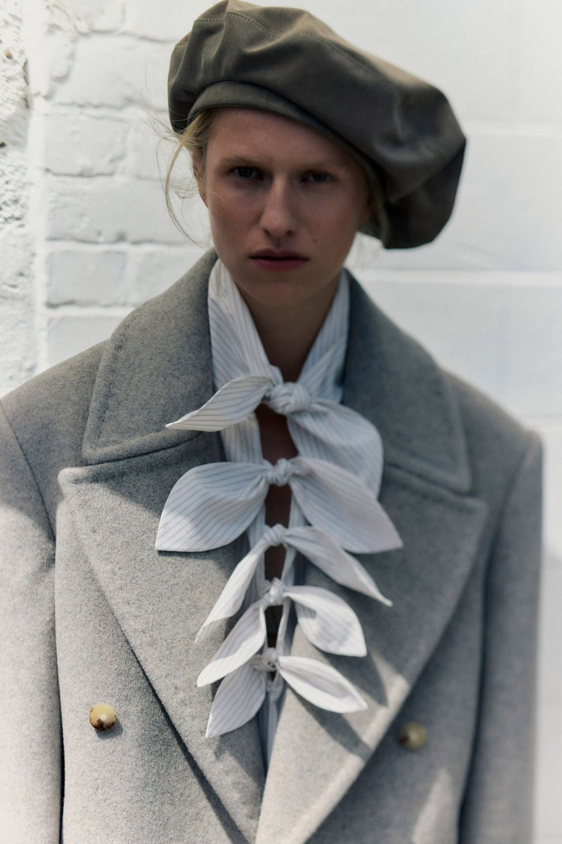 model wearing gray wool coat with sculptural white striped bow ties layered down the front with an oversized beret.