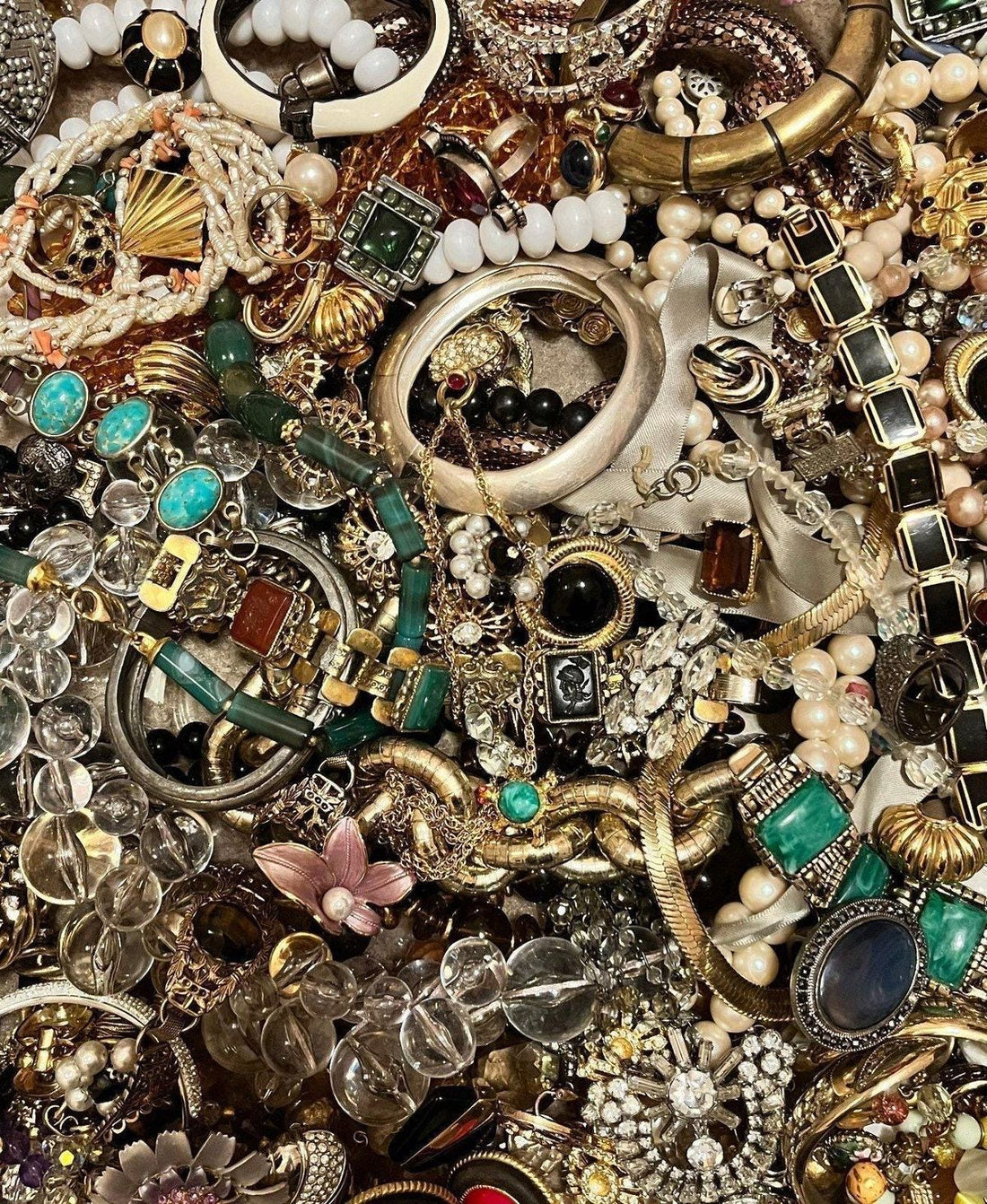 A lush overhead flat lay of vintage and costume jewelry — earrings, necklaces, bracelets, and bangles in gold, silver, turquoise, crystal, and pearl tumbled together.