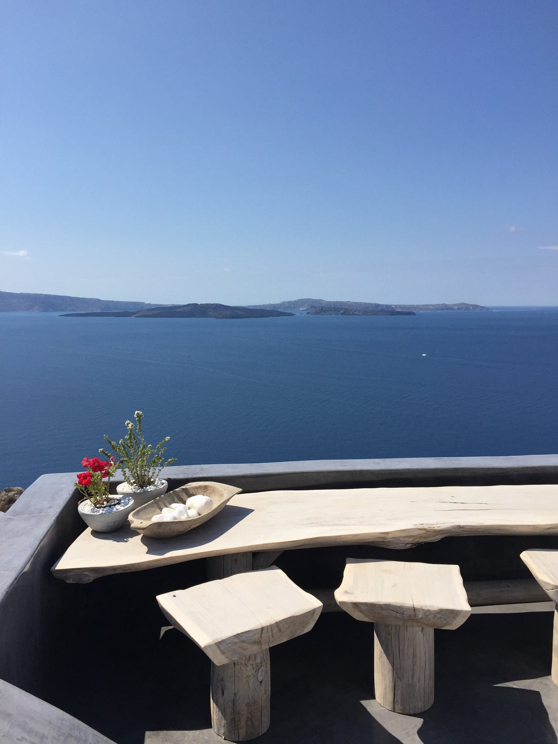 Ocean view terrace with minimal wooden seating overlooking deep blue water, evoking a calm, refined warm-weather travel atmosphere.