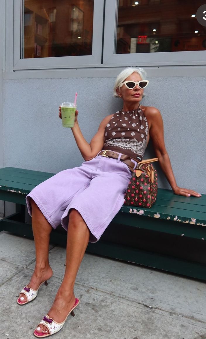 Mature woman slouching on a bench in lavender long denim jorts, louis vuitton and a matcha.